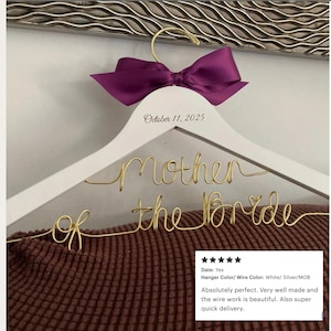 May include: A white wooden clothes hanger featuring a gold wire script that reads "mother of the bride" and a purple satin bow tied at the hook. The hanger is personalized with the printed date "October 11, 2025" on the upper wooden frame.