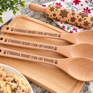 May include: Three light-colored wooden spoons with engraved text reading ETHERIDGE DESSERT COOK-OFF FINALIST, ETHERIDGE DESSERT COOK-OFF FAVORITE, and ETHERIDGE DESSERT COOK-OFF WINNER, each featuring a small cupcake icon at both ends of the text.