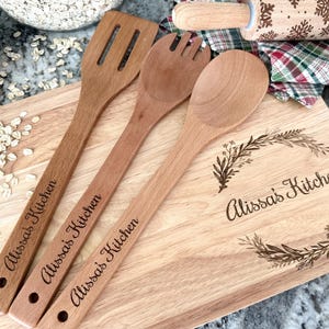 May include: Wooden kitchen utensils and a cutting board. The utensils include a slotted spatula, a fork, and a spoon. The cutting board is engraved with "Alissa's Kitchen" and floral designs. A rolling pin and a plaid cloth are in the background.