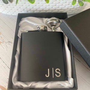 May include: A black metal flask with a silver funnel and the initials "J|S" engraved on the front. The flask is in a black box with a silver satin lining.