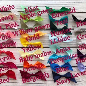 May include: A selection of satin bows in a range of colours, such as white, pink, yellow, and blue. Each bow is labelled with its colour name in a handwritten font. The bows are displayed on a light wood-grain surface.