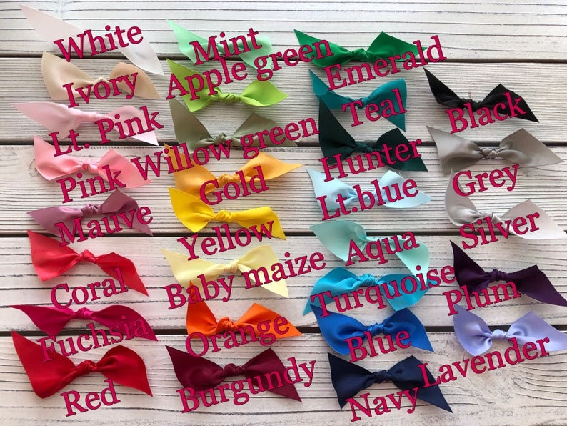 May include: A selection of satin bows in a range of colours, such as white, pink, yellow, and blue. Each bow is labelled with its colour name in a handwritten font. The bows are displayed on a light wood-grain surface.