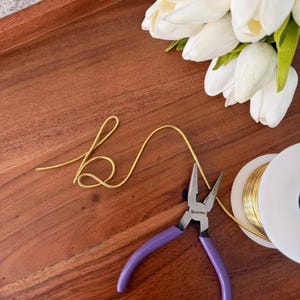 May include: A wooden tray holds a gold wire, purple-handled pliers, and a spool of gold wire. White tulips are arranged in the upper right corner. The gold wire is shaped into the letter "b".