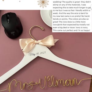 May include: A white wooden wedding dress hanger with a cream-colored bow and gold wire name. The date "May 10, 2025" is printed on the hanger. The wire spells out "Mrs. Vilman".