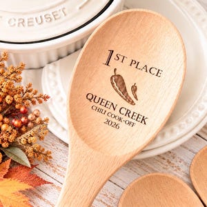 May include: A light-colored wooden spoon engraved with the text 1ST PLACE, QUEEN CREEK, CHILI COOK-OFF 2026, and an illustration of two chili peppers. The spoon has a smooth, rounded bowl and a long handle, showcasing the natural wood grain texture.
