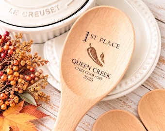 Puede incluir: Una cuchara de madera clara con un diseño grabado a láser que dice 1ST PLACE, QUEEN CREEK, CHILI COOK-OFF, 2026, acompañado de una ilustración de dos chiles centrada en la parte cóncava de la cuchara.