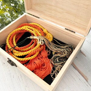 May include: A wooden box with a black interior, containing a set of graduation cords in orange, yellow, and green, with a gold tassel and a gold charm with the number "16" on it.
