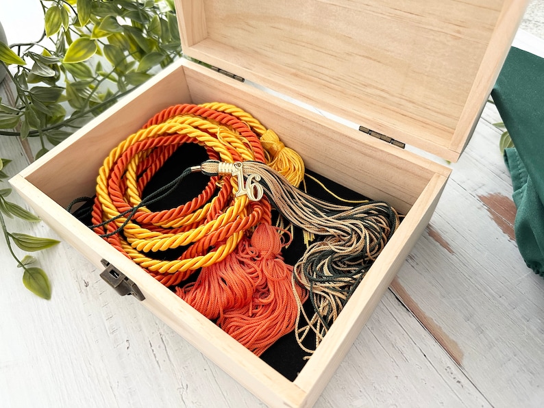 May include: A wooden box with a black interior, containing a set of graduation cords in orange, yellow, and green, with a gold tassel and a gold charm with the number "16" on it.