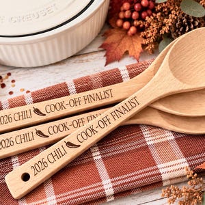 May include: Three light-colored wooden spoons with a smooth finish, each featuring a hole at the end of the handle. The handles are engraved with the text 2026 CHILI COOK-OFF FINALIST, accompanied by a small chili pepper icon between the words.