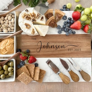 May include: A white and brown rectangular serving board with a marble top and a wooden bottom. The board is divided into two sections, one for cheese and one for other snacks. The board is decorated with the text "The Johnson Est. 2023".