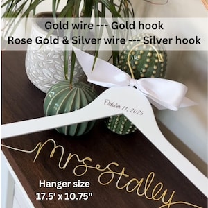 May include: A white wooden clothes hanger measuring 17.5 inches by 10.75 inches, featuring a gold wire script reading mrs staley, a gold hook, and a white satin ribbon bow. The text October 11, 2025 is printed on the hanger frame.