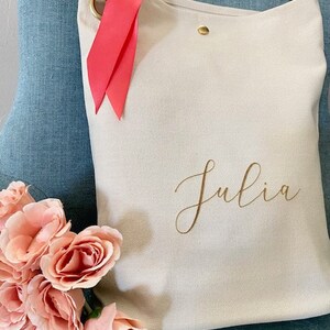 May include: A cream canvas tote bag with a brown leather strap and gold-coloured hardware. The bag features the name "Julia" in gold script. A bright pink ribbon is tied to the strap. A bouquet of pink roses is in the foreground.