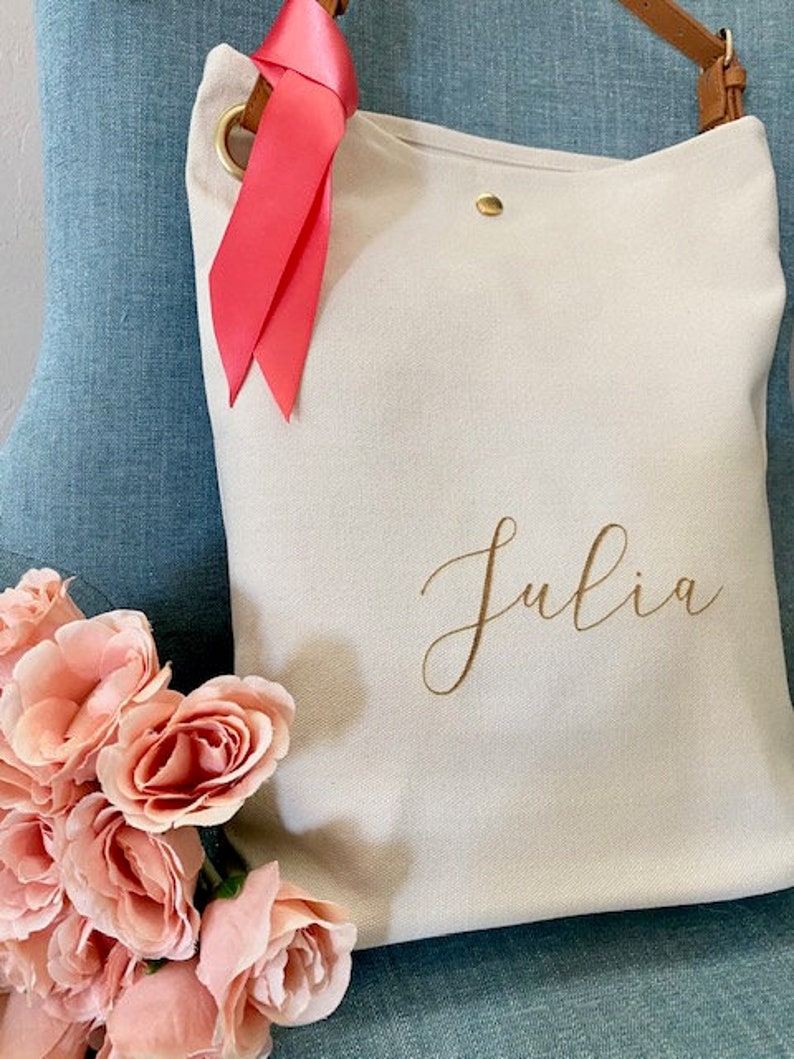 May include: A cream canvas tote bag with a brown leather strap and gold-coloured hardware. The bag features the name "Julia" in gold script. A bright pink ribbon is tied to the strap. A bouquet of pink roses is in the foreground.