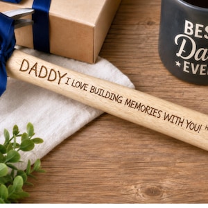 May include: A wooden hammer with the words "DADDY I LOVE BUILDING MEMORIES WITH YOU! Happy Father's Day! Love Josh" engraved on the handle. A blue ribbon is tied around the hammer head. A mug that says "BEST Dad EVER" is also in the image.