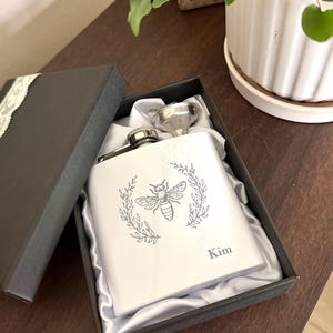 May include: A white stainless steel flask with a bee and laurel wreath design, presented in a black gift box. The flask includes a silver funnel and is personalized with the name "Kim". The box is lined with satin.