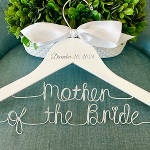 May include: A white wooden clothes hanger featuring a silver wire script that reads Mother of the Bride. The hanger is adorned with a white satin ribbon bow at the top and includes the printed text December 20, 2024, on the wooden frame.