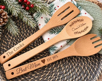 May include: Three light-colored wooden kitchen utensils including a slotted spatula engraved with The Cooks, a spoon engraved with Owen's Kitchen, and a salad fork engraved with Best Mom heart EST 1975, each featuring a hole in the handle.