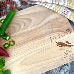 May include: A wooden cutting board with the text "1st Place" and a chili pepper graphic, along with "AEW Chili Cook-Off 2025" engraved on the surface. Green jalapeños, red tomatoes, and cilantro are arranged on the board.