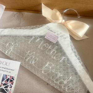 May include: A white wooden clothes hanger wrapped in clear bubble wrap, featuring a cream-colored satin ribbon bow at the top. The hanger is printed with the text "Mother of the Bride" and the date "November 30, 2026" in a cursive script font.