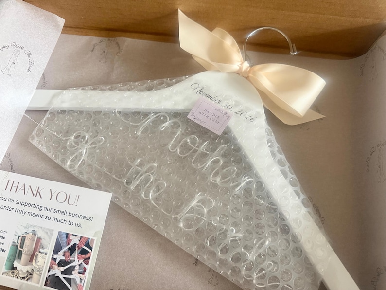 May include: A white wooden clothes hanger wrapped in clear bubble wrap, featuring a cream-colored satin ribbon bow at the top. The hanger is printed with the text "Mother of the Bride" and the date "November 30, 2026" in a cursive script font.
