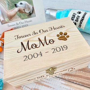May include: A wooden box with a hinged lid and a latch. The box is engraved with the words "Forever In Our Hearts MoMo 2004 - 2019" and a paw print. The box is surrounded by a dog toy, a photo frame, and a plush dog toy shaped like a bottle of wine.