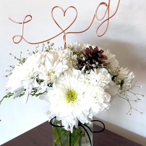 May include: A white flower arrangement in a clear glass jar with a black ribbon tied around the top. A copper wire sculpture of the letters "L" and "G" with a heart in between is placed in the arrangement.