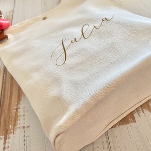 May include: Cream canvas tote bag with the name "Julia" in gold script. It features a brown leather strap and a red ribbon tied to a metal ring. The bag is on a light-coloured wooden surface.