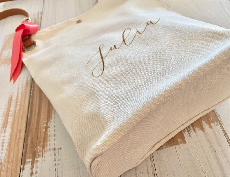 May include: Cream canvas tote bag with the name "Julia" in gold script. It features a brown leather strap and a red ribbon tied to a metal ring. The bag is on a light-coloured wooden surface.