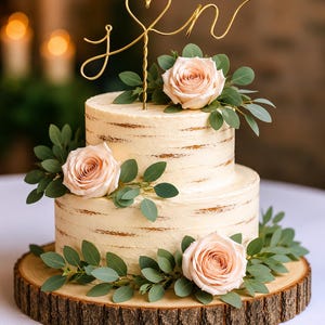 May include: Two-tiered wedding cake with cream frosting, decorated with peach-colored roses and green leaves. A gold wire cake topper with a heart and initials sits atop the cake. The cake rests on a wooden cake stand.