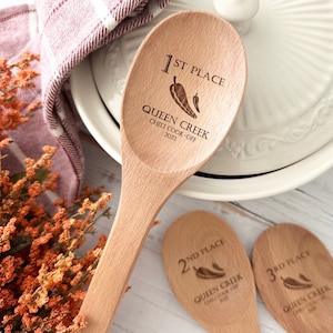 May include: Wooden spoons engraved with "1st Place," "2nd Place," and "3rd Place" for the Queen Creek Chili Cook-Off 2022. Each spoon features a chili pepper graphic. The spoons are light brown and rest on a white surface.