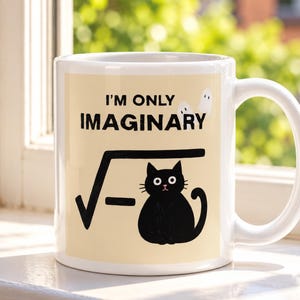 May include: A white ceramic mug featuring a light beige rectangular graphic with the black text I'M ONLY IMAGINARY above a square root symbol containing a negative sign and a black cat illustration with two small white ghost figures nearby.