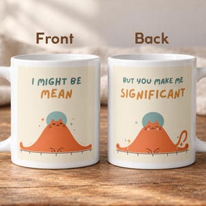 May include: Two white ceramic mugs featuring an orange cat illustration. The left mug displays the text I MIGHT BE MEAN above the cat. The right mug displays the text BUT YOU MAKE ME SIGNIFICANT above the cat. Both mugs show a cat with a blue halo.