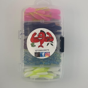 May include: A clear plastic tackle box containing five compartments of soft plastic fishing lures in pink, dark blue, purple, blue-speckled, and white-green colors. A circular sticker features a red muscular fish logo and the text @TONKABAITS.