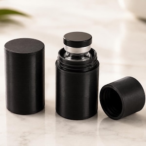 May include: Three cylindrical black metal containers with matte finishes. One container is fully closed, one is open with a small glass vial inside, and one is a separate screw-on cap. The containers have a smooth, uniform texture and design.