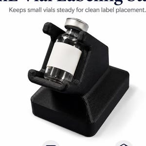 May include: A black 3 mL Vial Labeling Stand with a rectangular base and a cradle for holding a small glass vial with a silver cap and a blank white label. The text 3 mL Vial Labeling Stand, Keeps small vials steady for clean label placement, Steady Hold, Easy Labeling, and Compact Design are visible.