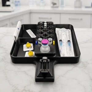 Pode incluir: Uma bandeja organizadora médica de plástico preto com múltiplos compartimentos para suprimentos. Contém duas seringas embaladas, vários frascos de vidro pequenos com tampas coloridas, dois itens amarelos e um pequeno pacote de papéis brancos.