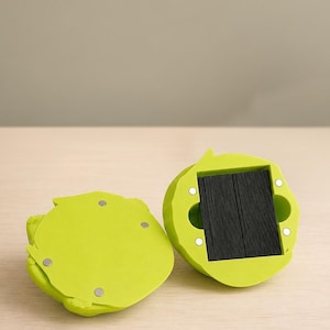Könnte beinhalten: Zwei limettengrüne, blattförmige Magnethalter. Einer zeigt die Unterseite mit vier Magneten, der andere einen dunklen, rechteckigen Innenraum. Die Halter liegen auf einer hellen Holzoberfläche.