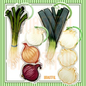 May include: A collection of four acrylic keychains featuring illustrations of vegetables. The designs include a green leek, a stack of white, yellow, and red onions, a dark green leek, and a stack of three white garlic bulbs, all with light green rings.