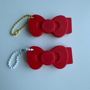 May include: Two red bow-shaped keyrings. One has a gold ball chain, the other a white beaded chain. The bows have a textured surface and a circular centre. These are likely accessories or charms.