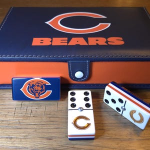 May include: A navy blue and orange Chicago Bears domino set. The set includes a box with the Bears logo and the word "BEARS". Several dominoes are visible, featuring the team's logo and colors.