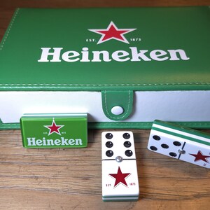 Op de afbeelding: Een groene Heineken-doos met een rood sterlogo, een kleine groene doos en twee dominostenen. De dominostenen zijn wit met zwarte stippen en een rode ster. De doos is gesloten met een witte knop.