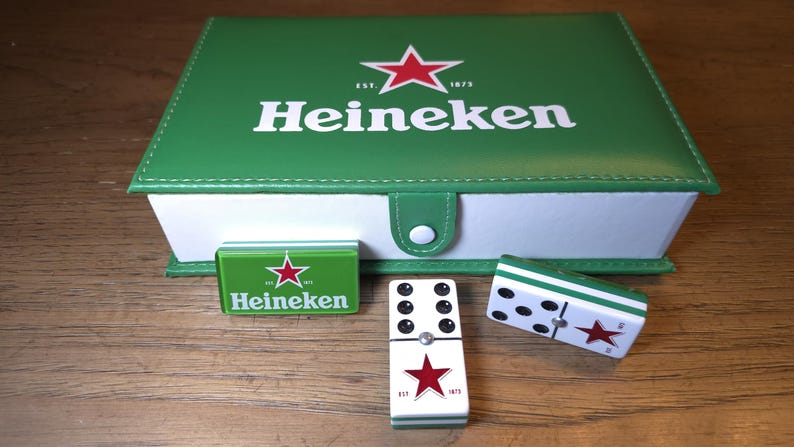 Op de afbeelding: Een groene Heineken-doos met een rood sterlogo, een kleine groene doos en twee dominostenen. De dominostenen zijn wit met zwarte stippen en een rode ster. De doos is gesloten met een witte knop.