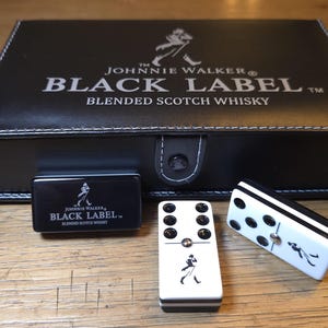 May include: A black leather box set with the Johnnie Walker Black Label logo, a small black box, and two dominoes. The dominoes are white with black dots and a Johnnie Walker logo.