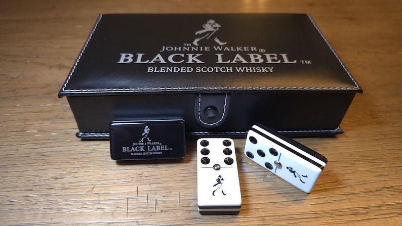 May include: A black leather box set with the Johnnie Walker Black Label logo, a small black box, and two dominoes. The dominoes are white with black dots and a Johnnie Walker logo.