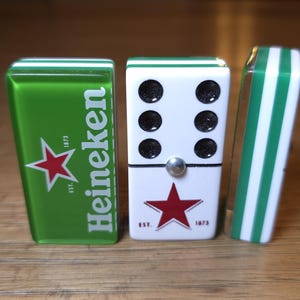 Op de afbeelding: Dominoset met een groen en wit thema. E&eacute;n domino heeft het Heineken-logo met een rode ster. De middelste domino is wit met zwarte stippen en een rode ster. De derde domino heeft groen-witte strepen.