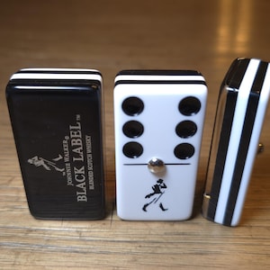 May include: Three dominoes with a black and white design. One domino is black with white stripes and the text "BLACK LABEL" and a logo. Another domino is white with black dots and a logo. The third domino is black and white striped.