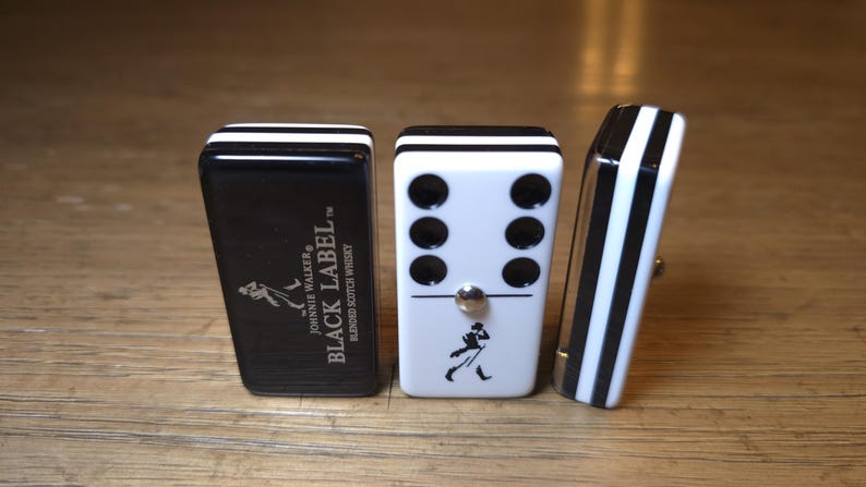 May include: Three dominoes with a black and white design. One domino is black with white stripes and the text "BLACK LABEL" and a logo. Another domino is white with black dots and a logo. The third domino is black and white striped.