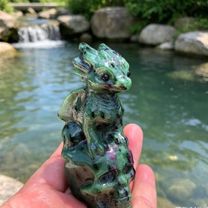 May include: A carved stone figurine featuring a small green dragon perched atop a human skull. The sculpture is crafted from a polished mineral with deep green hues and distinct dark and magenta-colored inclusions throughout the textured surface.
