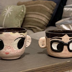 Puede incluir: Dos tazas de cerámica con forma de cabezas humanas y orejas prominentes. La taza de la izquierda muestra un rostro pálido con ojos grandes, pecas y labios rojos. La de la derecha tiene un rostro bronceado, cabello negro y gafas de montura negra.