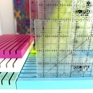 May include: A stack of four rectangular plastic organisers with parallel slots in magenta, white, mint green, and light blue. A clear Olfa QR-6S acrylic quilting ruler is placed vertically against the stack, showing a height of 10 cm.